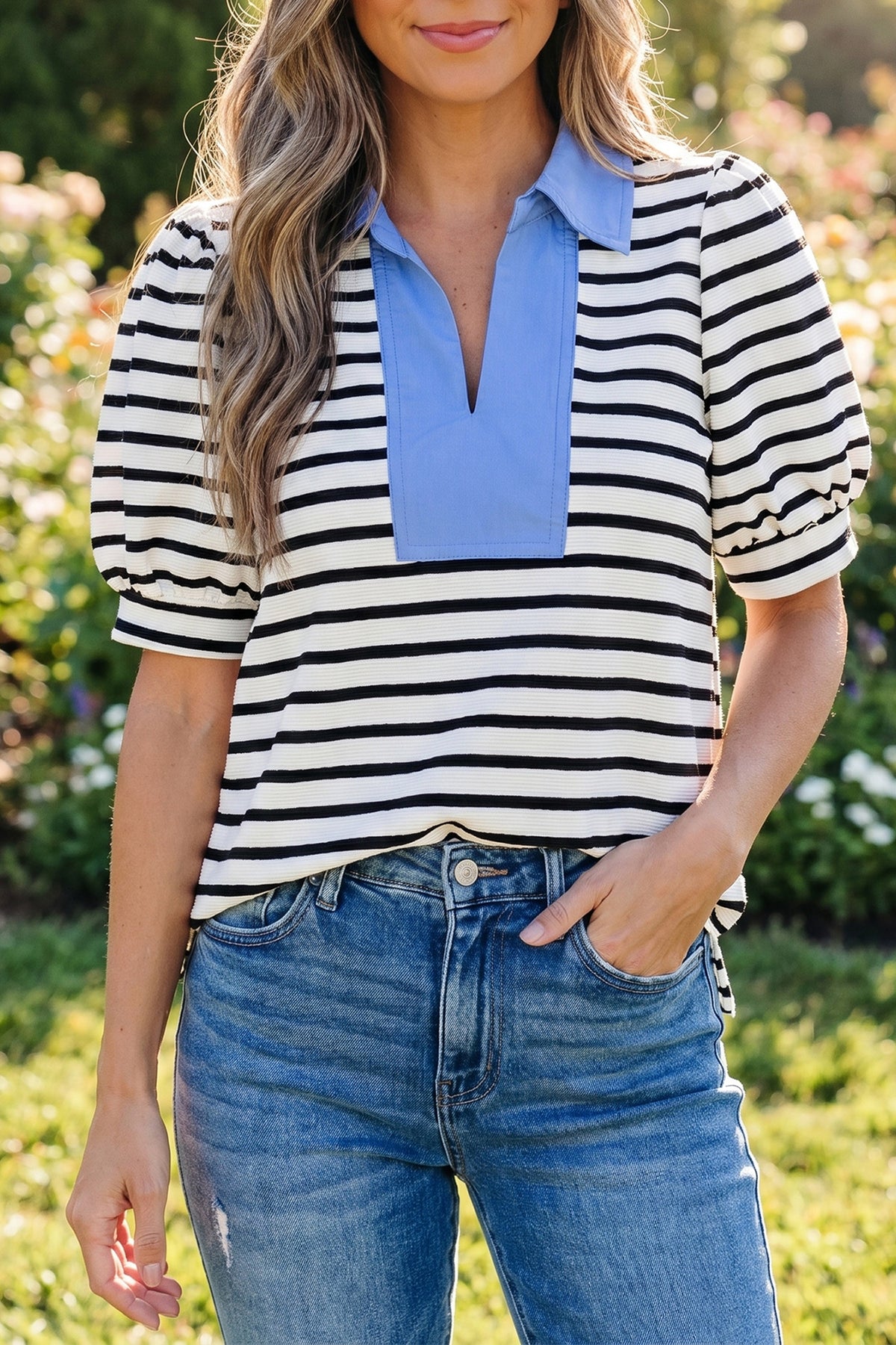 Woman wearing black and white striped short puff sleeve blouse with blue color block polo collar, standing outdoors in jeans
