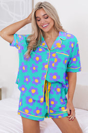Woman wearing teal short sleeve pajama set with purple and yellow flower print, sitting on bed