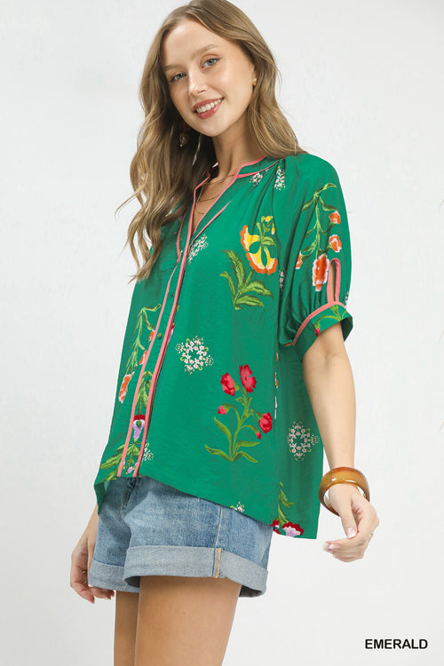 Woman wearing emerald botanical print blouse with pink contrast trim, button-up, short sleeves, and denim shorts