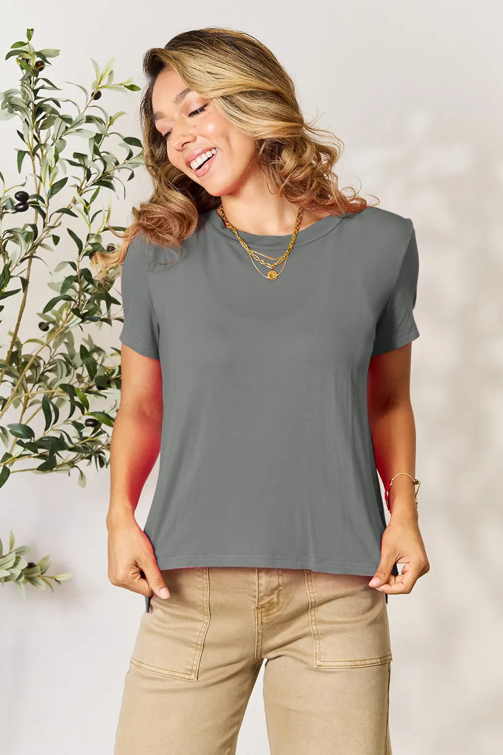 Woman wearing grey round neck short sleeve t-shirt with beige pants, smiling near olive branch