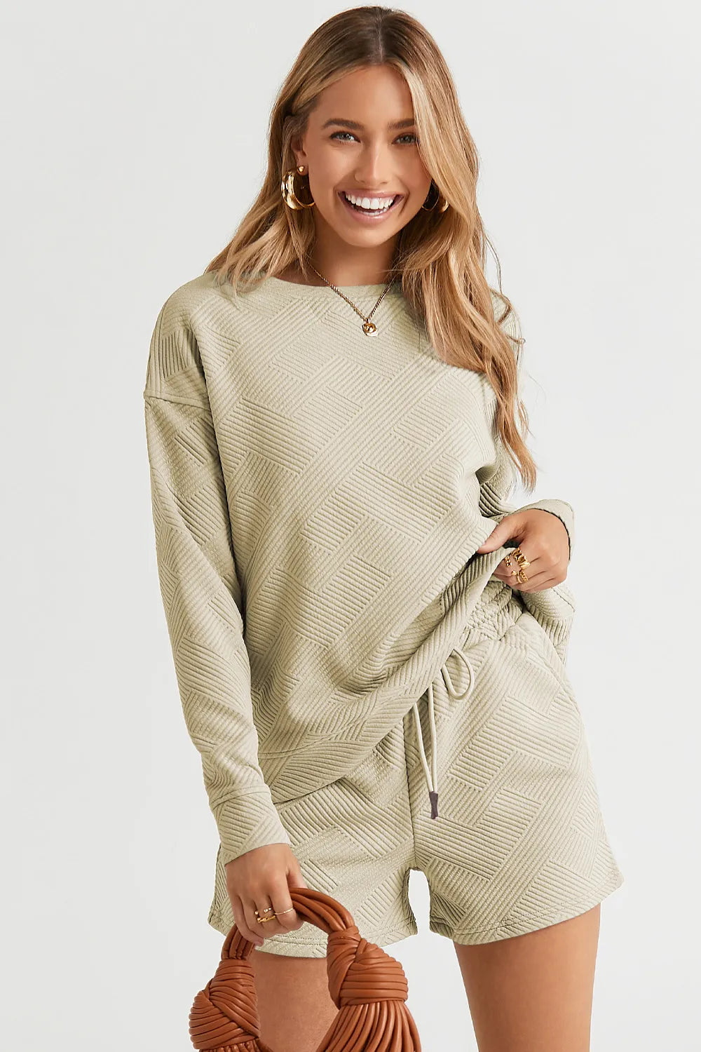Model wearing olive green textured long sleeve top and matching drawstring shorts, holding a brown woven bag